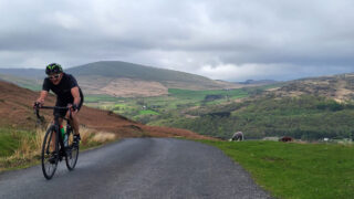 Adrian is pictured on a bicycle on. a road, having just ridden up what looks like a large hill. There is a beautiful landscape and hills in the background.